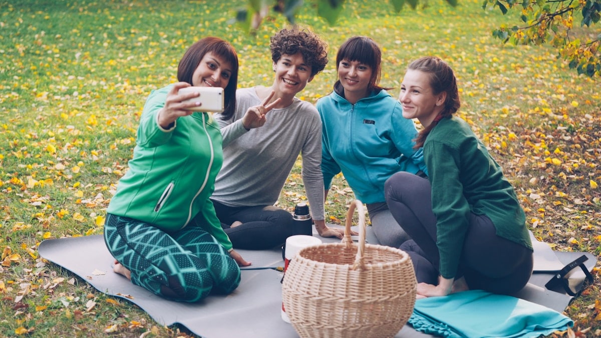 Pretty young women are taking selfie with smartphone during picnic in park sitting on grass, posing and laughing having fun. Nature, friendship and modern technology concept.